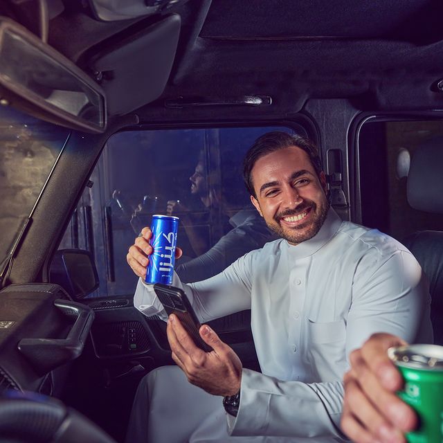 A man in a car smiling while holding a Kinza Cola can